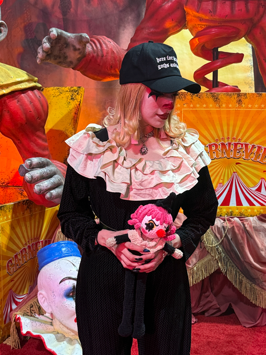 A person stands on a red carpet in front of a Halloween-themed clown backdrop, wearing a black outfit, black cap, and holding a pink plush toy.