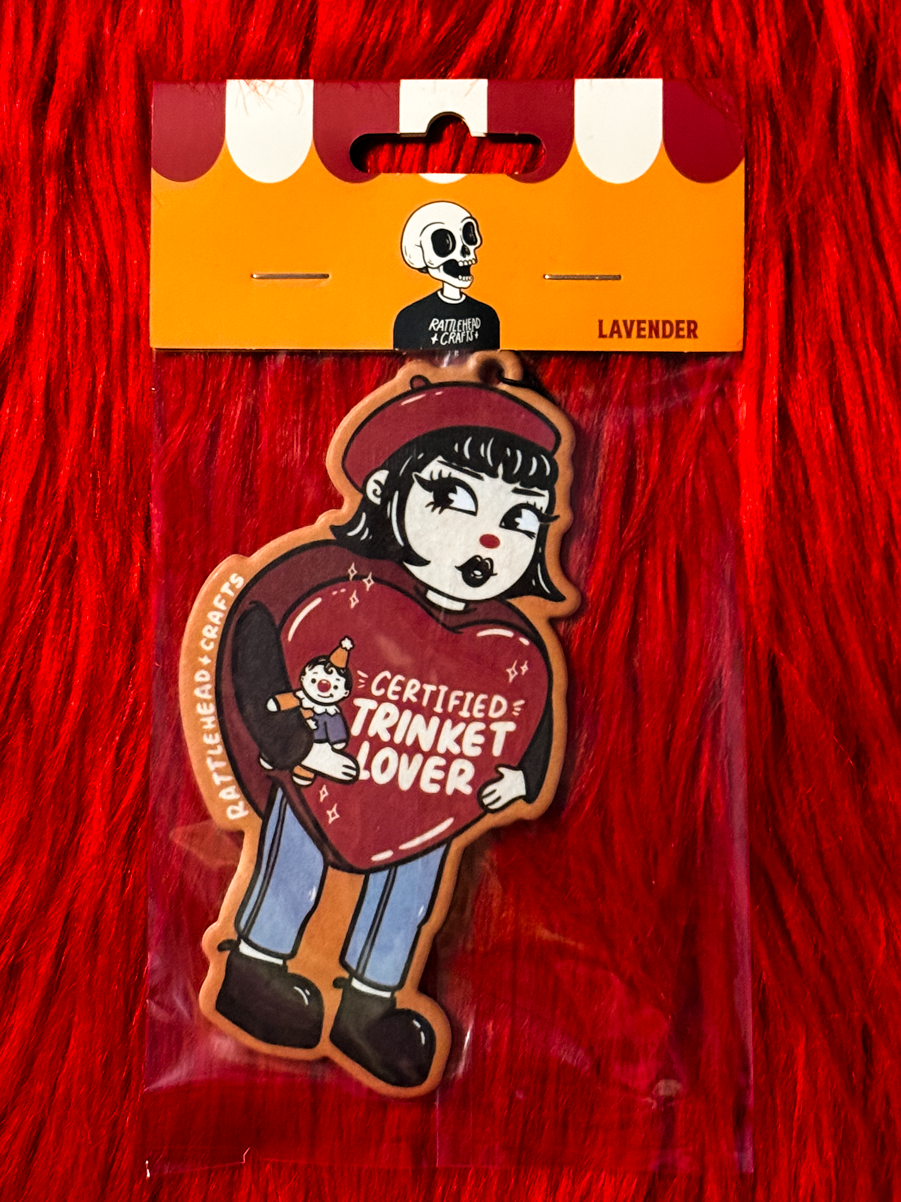 A packaged air freshener shaped like a girl holding a red heart that reads 'CERTIFIED TRINKET LOVER,' shown on a bright red furry background. The card header says LAVENDER and includes a small skull graphic.