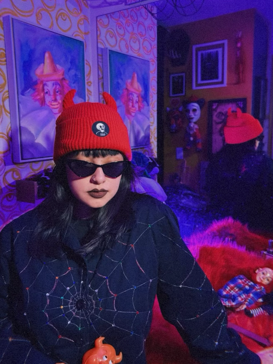 Person wearing a red beanie with devil horns and dark sunglasses in a colorful room with artwork on the walls.